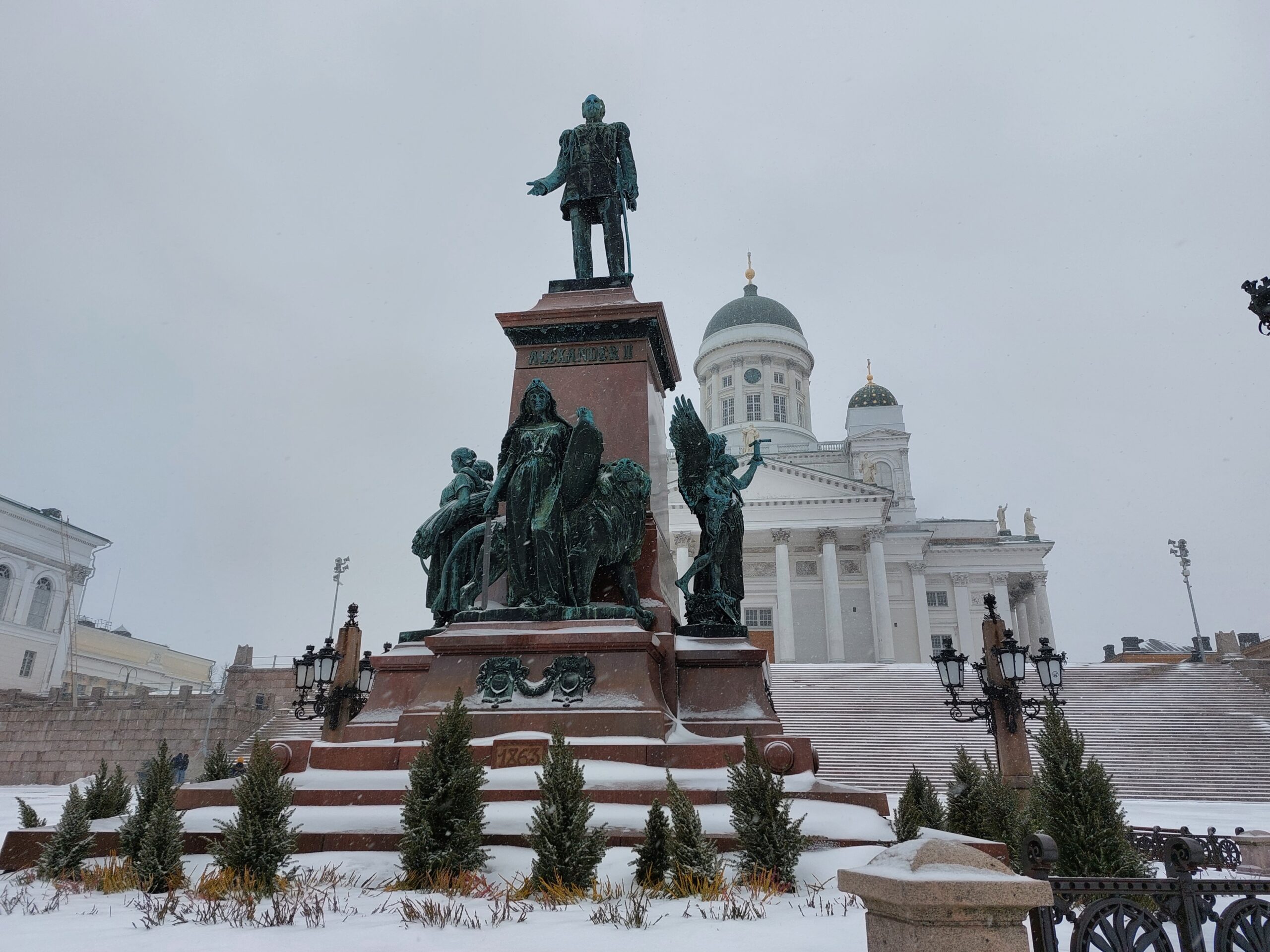 Denkmal in Helsinki mit Statue und Skulpturen vor einer weißen Kirche im Winter, bedeckt mit Schnee.