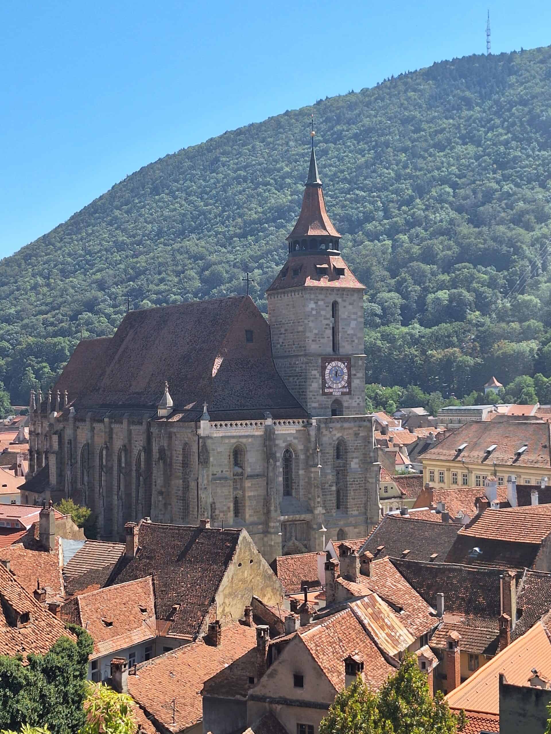 Alte Kirche mit steilem Turm vor grüner Bergkulisse, umgeben von roten Ziegeldächern. in Brasov einer mittelalterlichen Stadt in Transsylvanien - Rumänien